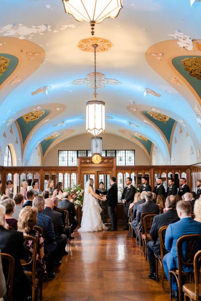 Bride and groom exchange vows in ornate venue with guests seated, highlighting elegant architecture.