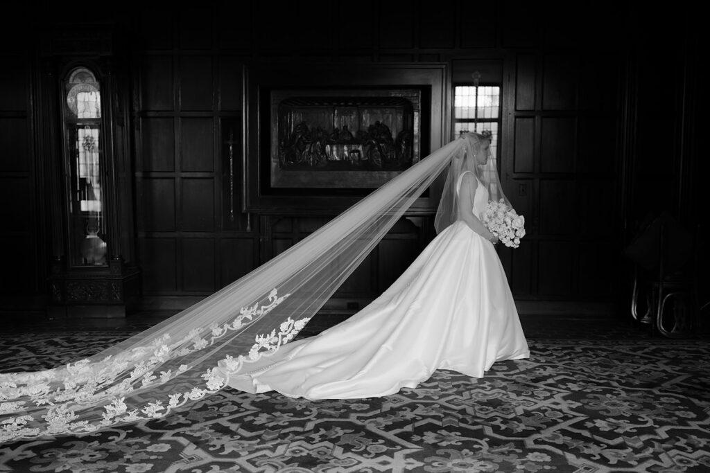 Elegant bride in flowing gown and long veil walking indoors, holding bouquet of flowers. Classic black and white photo.