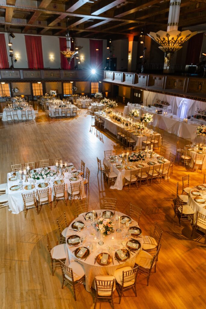 Elegant ballroom wedding reception setup with round tables, white linens, and floral centerpieces under warm lighting.
