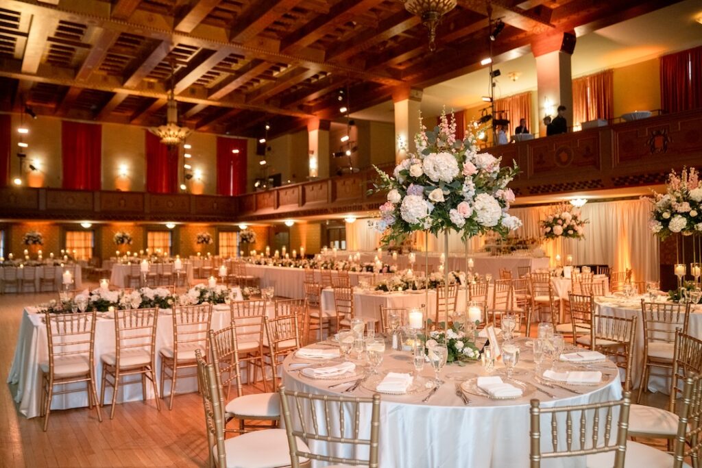 Elegant wedding reception hall with round tables, floral centerpieces, and golden chairs under a wooden ceiling.