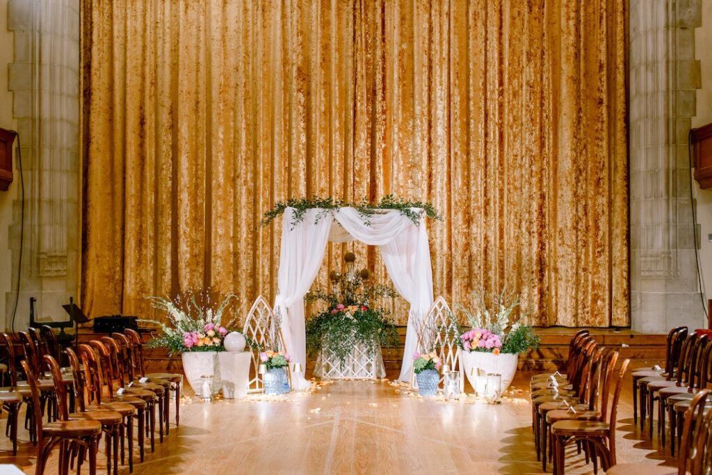 Elegant indoor wedding venue with floral decor, white draped arch, and golden backdrop curtain.