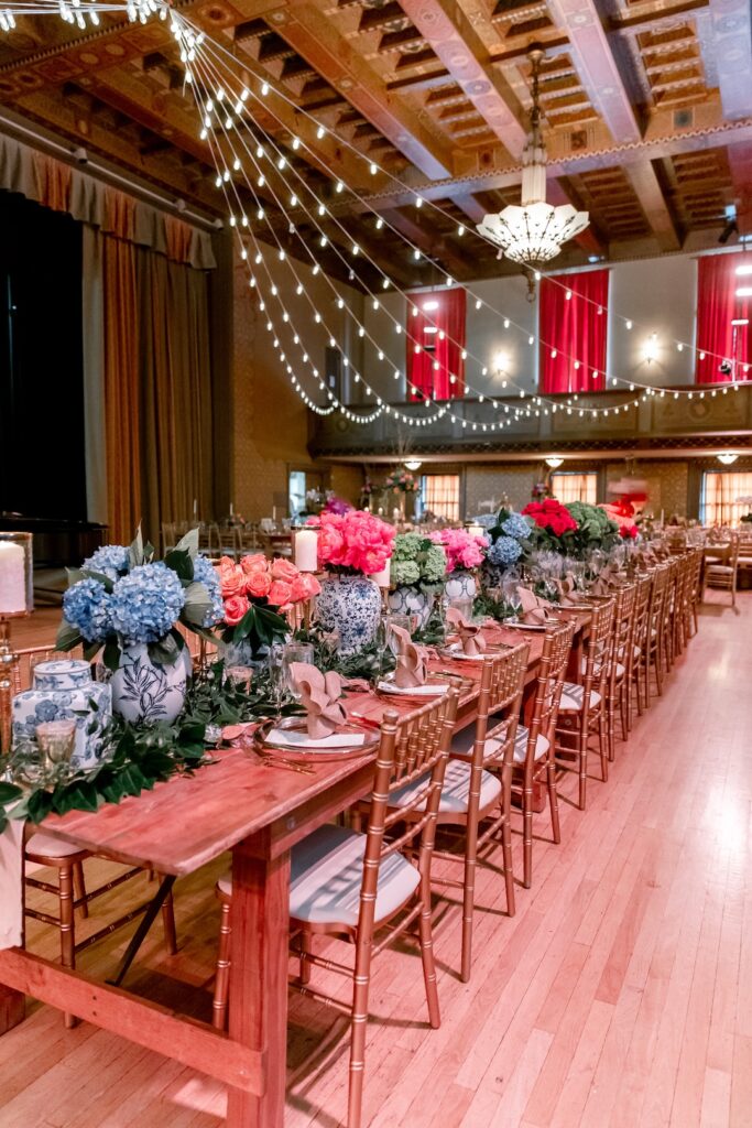 Elegant event setup with floral centerpieces, string lights, and wooden tables in a decorated ballroom.