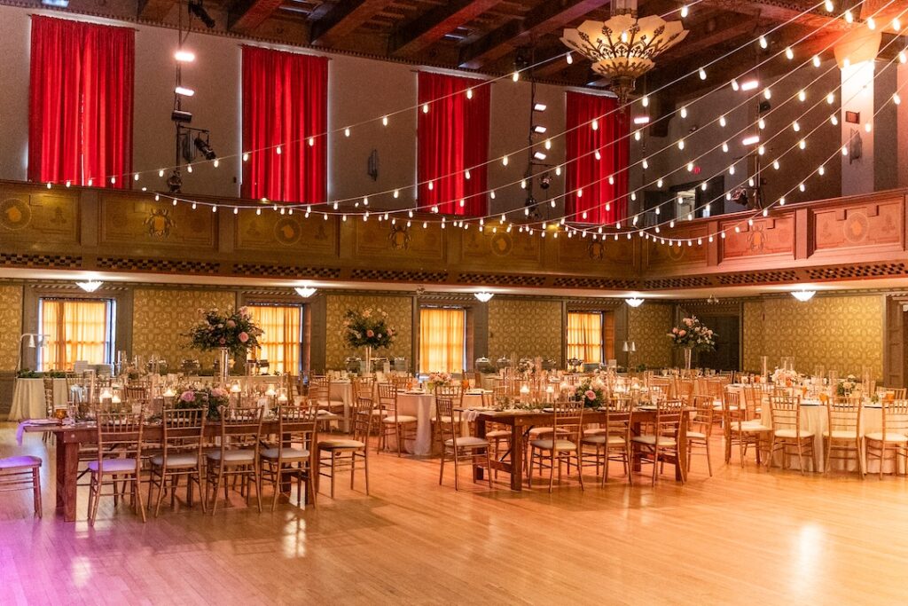 Elegant ballroom with string lights and decorated tables set for an event, featuring red drapes and warm lighting.