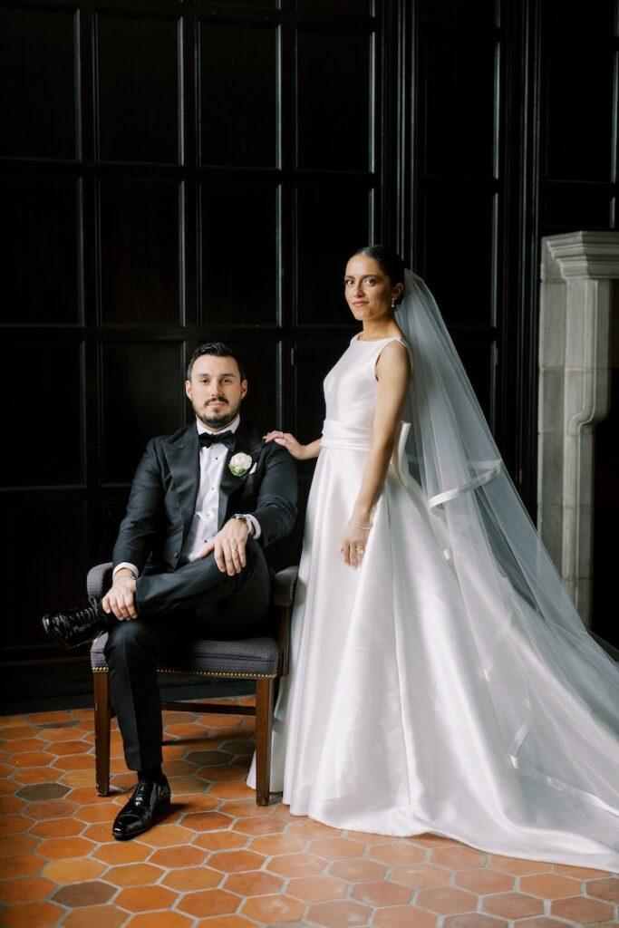 Bride and groom in elegant attire, wedding portrait in formal setting with dark wood backdrop.