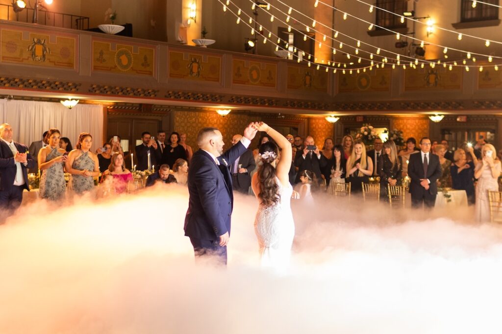 Bride and groom dance in smoke under string lights at a romantic wedding reception surrounded by guests.