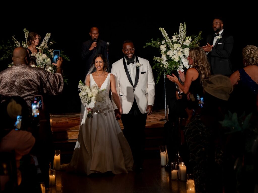 Bride and groom smiling, walk down the aisle celebrating their wedding, surrounded by guests and flowers.