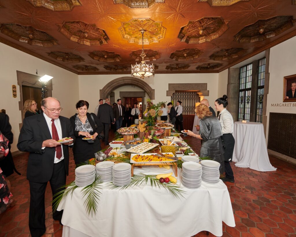 Elegant buffet setup at a formal event with guests serving food, ornate ceiling, and stylish decor.