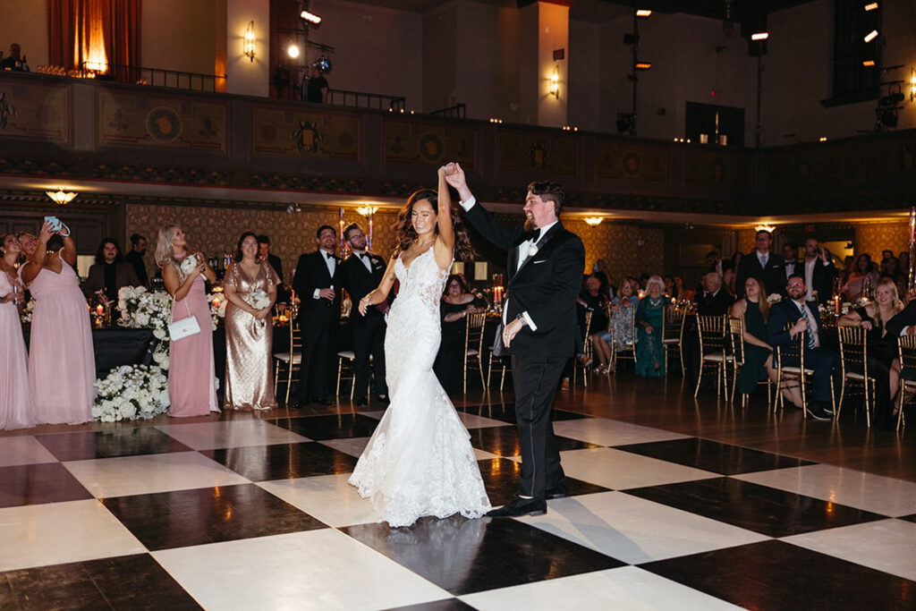 Bride and groom dance at wedding reception, guests in formal attire watch and take photos.
