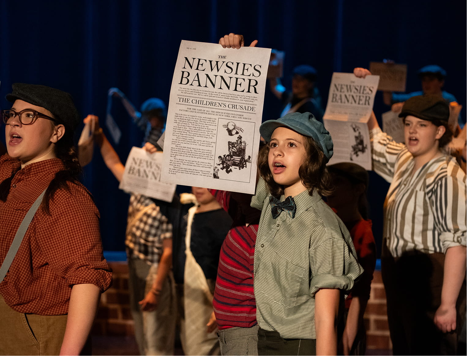 Children in vintage costumes perform Newsies on stage, holding newspapers, singing with enthusiasm under stage lights.
