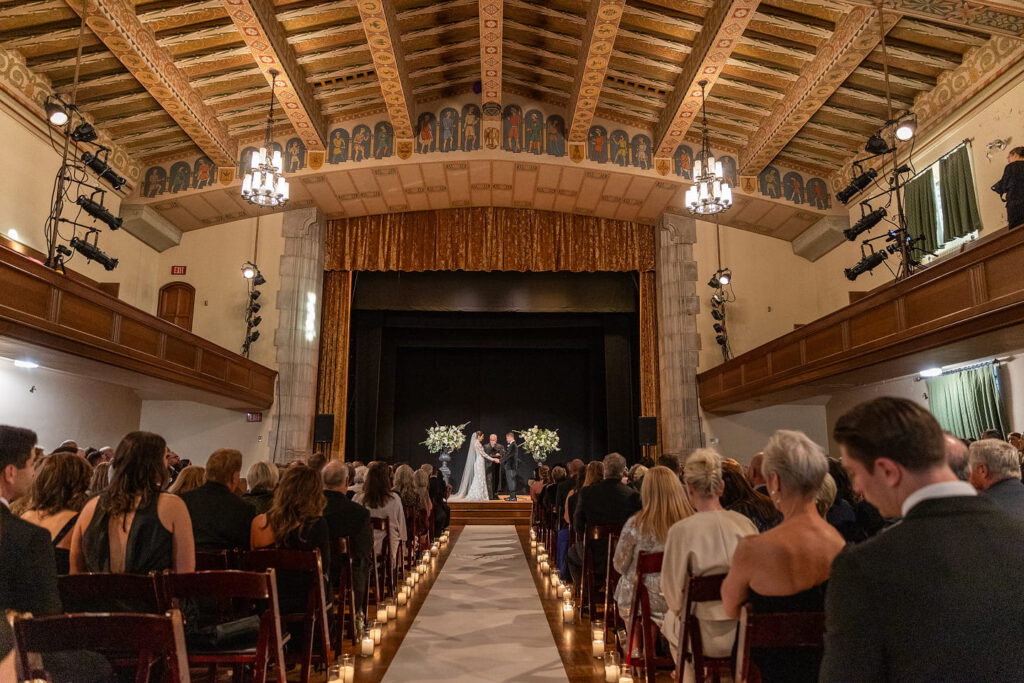 Elegant indoor wedding ceremony in a grand hall with guests seated, chandeliers, and ornate ceiling.