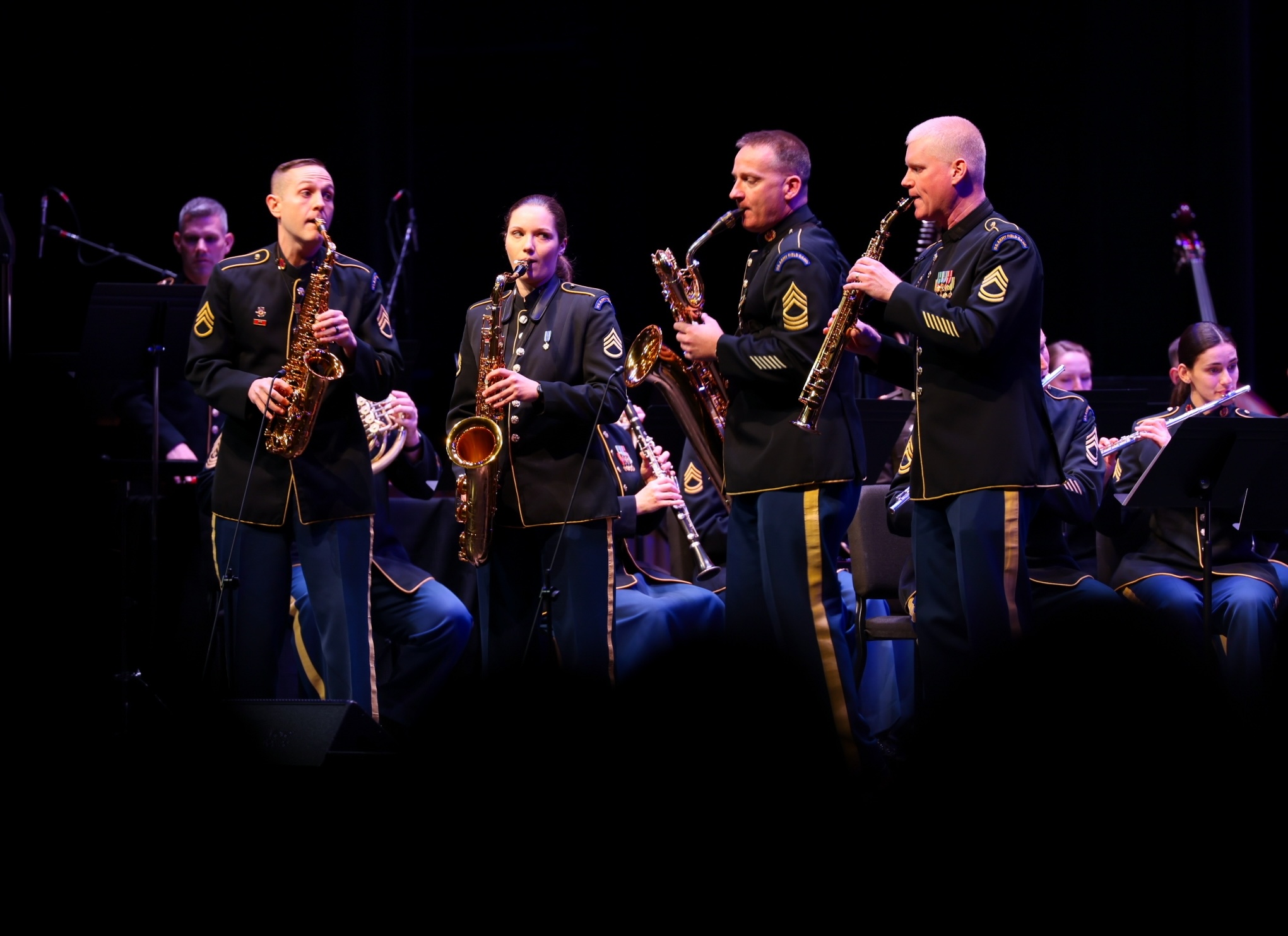 Military band musicians performing onstage with saxophones and woodwind instruments, wearing formal uniforms.