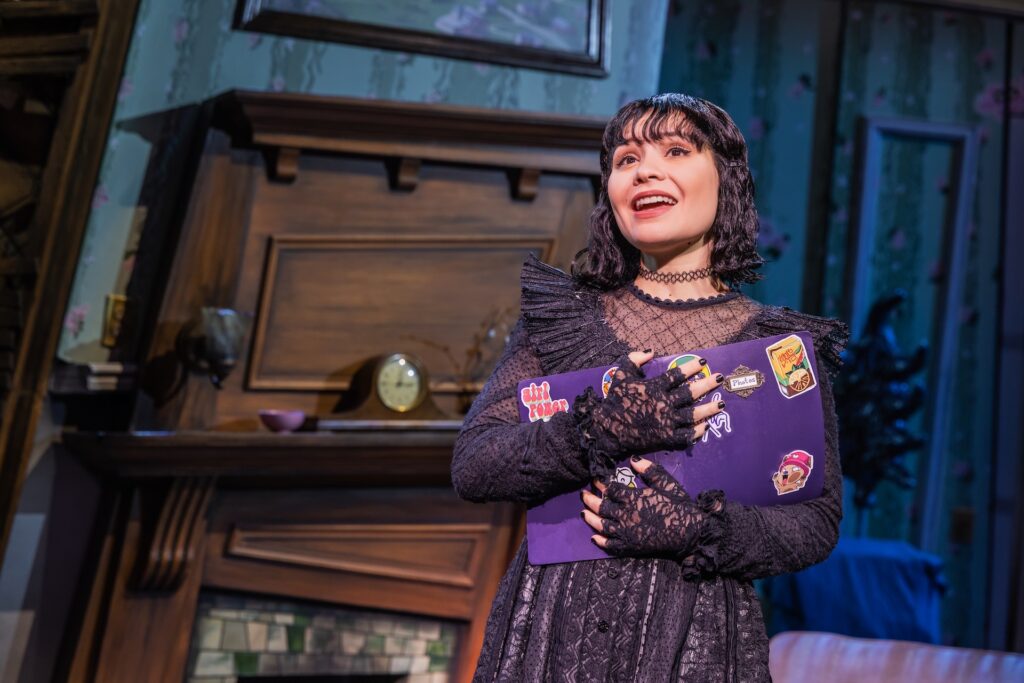 Actress in lace dress holding a sticker-covered notebook onstage, with vintage decor background.