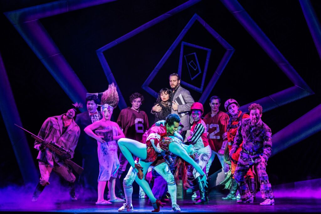 Vibrant musical theater cast in colorful costumes performing energetically on stage with dramatic lighting.