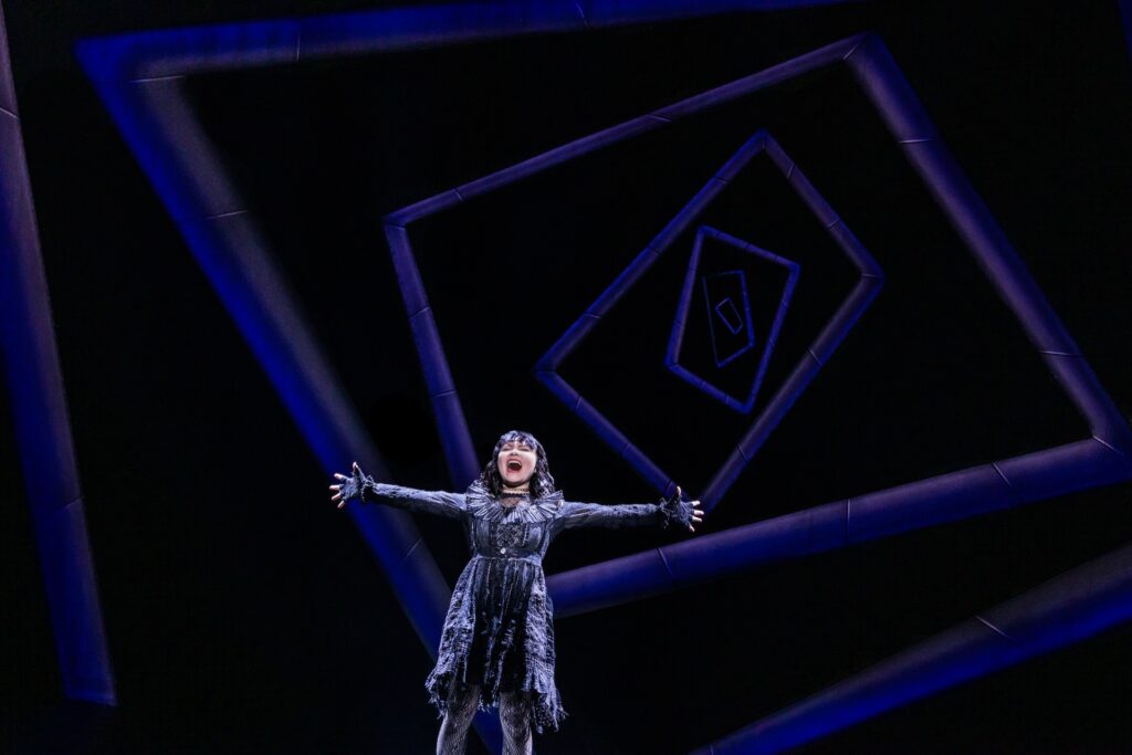 Performer in intricate costume on stage with geometric blue backdrop, arms outstretched in dramatic pose.