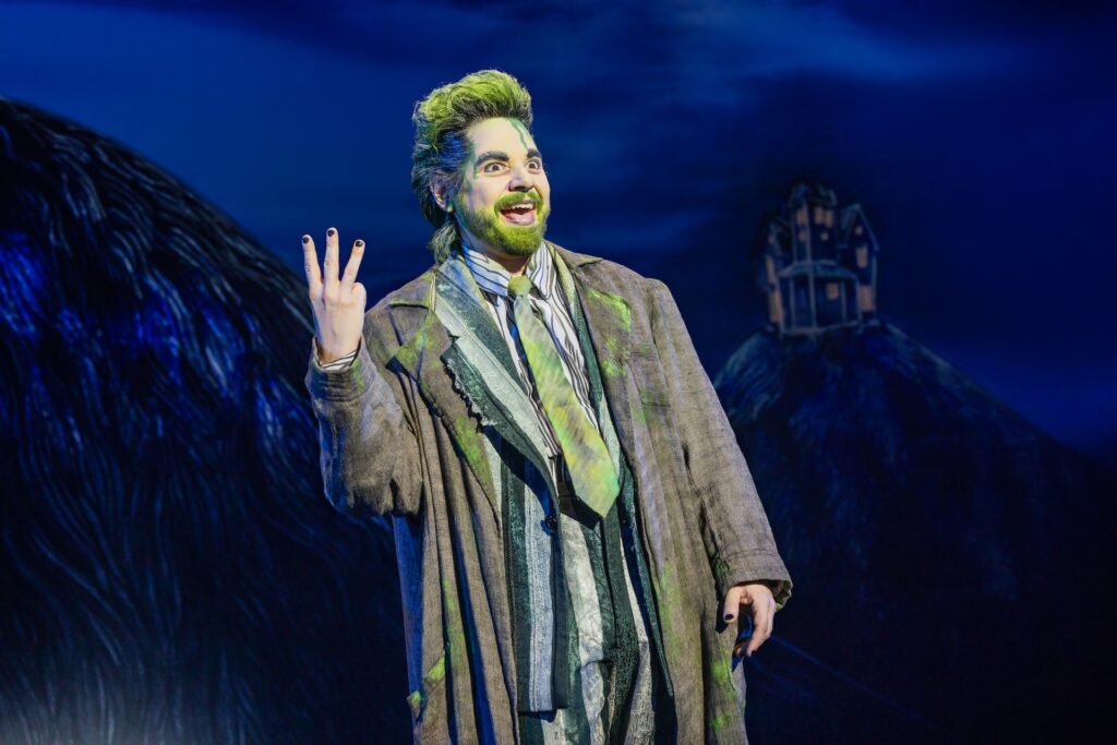 Character with green hair and suit performing energetically on stage, dark scenic backdrop with a house on a hill.