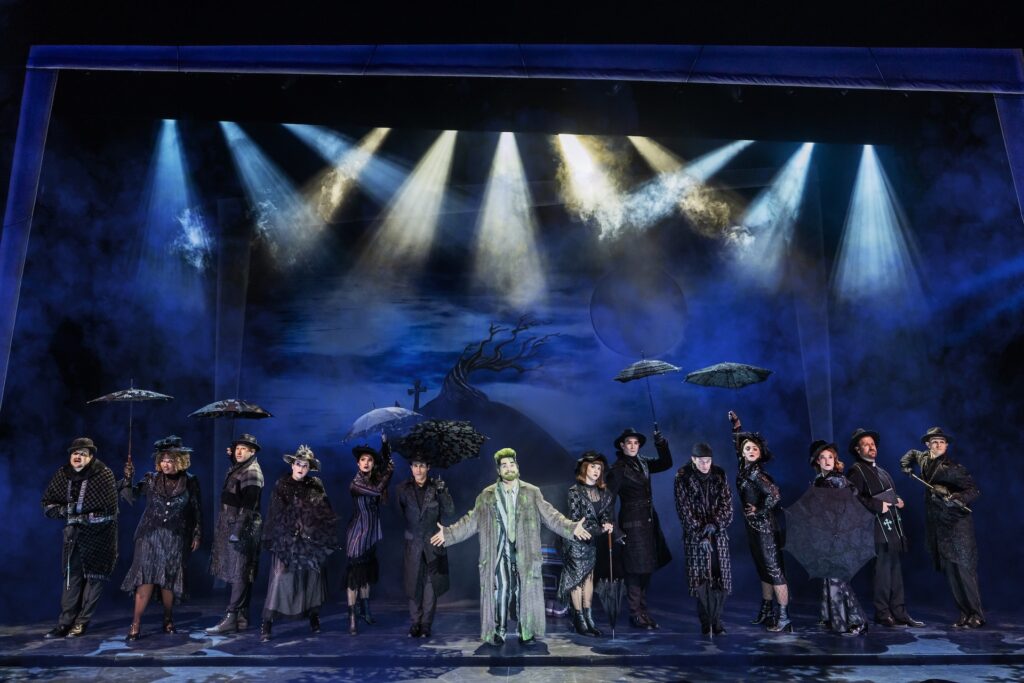 Theatrical cast in dark costumes with umbrellas on stage under dramatic blue lighting, performing a musical scene.