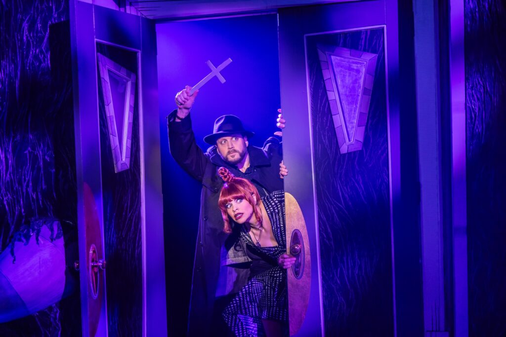 Two people in a theatrical scene; man holding a cross, woman peeking, dramatic purple lighting.