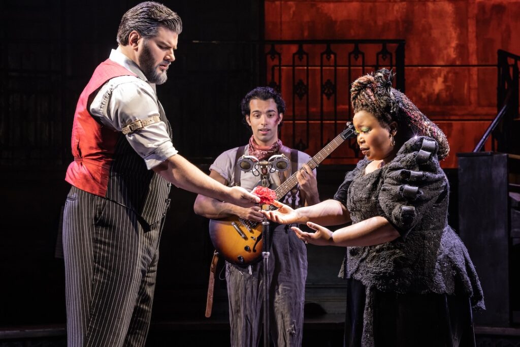 Stage performance with actors exchanging a flower, musician holding a guitar in the background, dramatic setting.