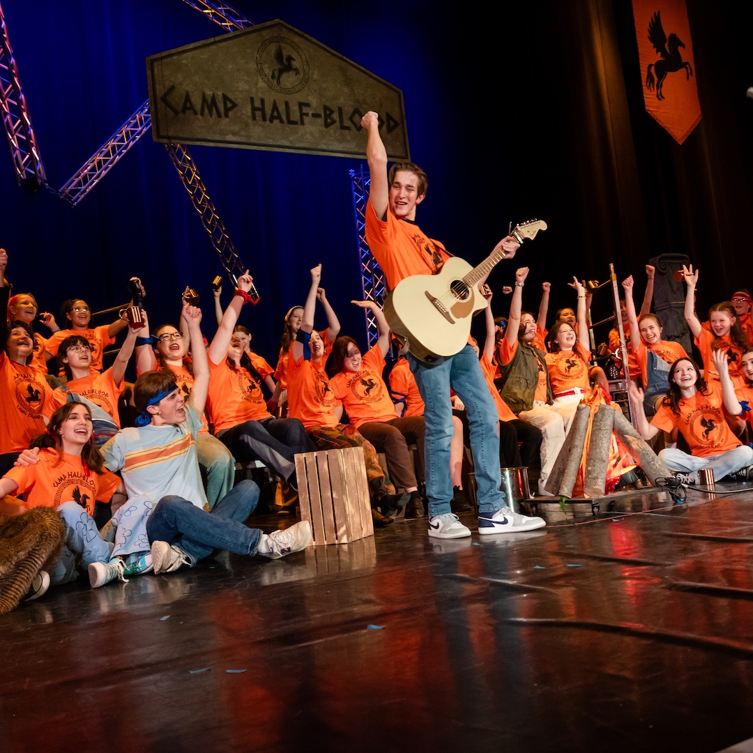 Group on stage at Camp Half-Blood gathering, cheering with guitar, orange shirts, and a campfire setup.