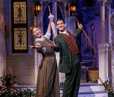 Two actors in vintage costumes perform joyfully on stage with props, in front of a decorated porch setting.