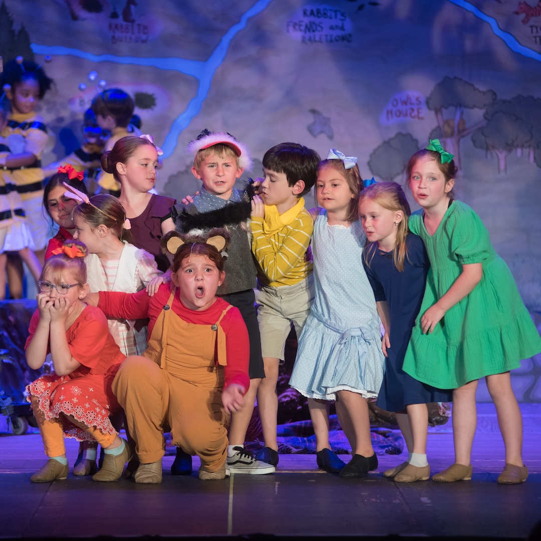 Children performing in colorful costumes on stage during a lively play with a forest backdrop.